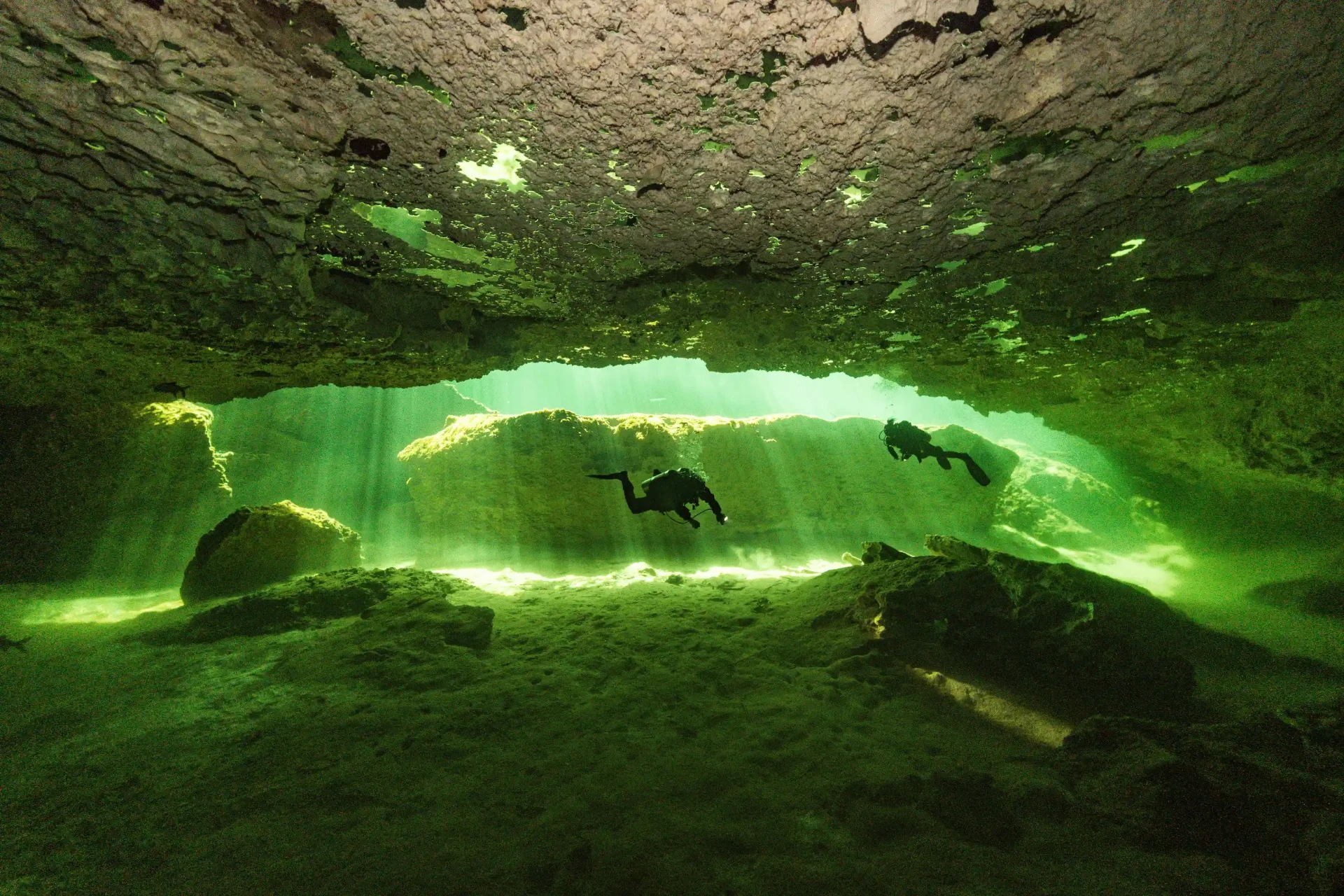 Two scuba divers silhouetted against bright green water below a rocky cenote overhang