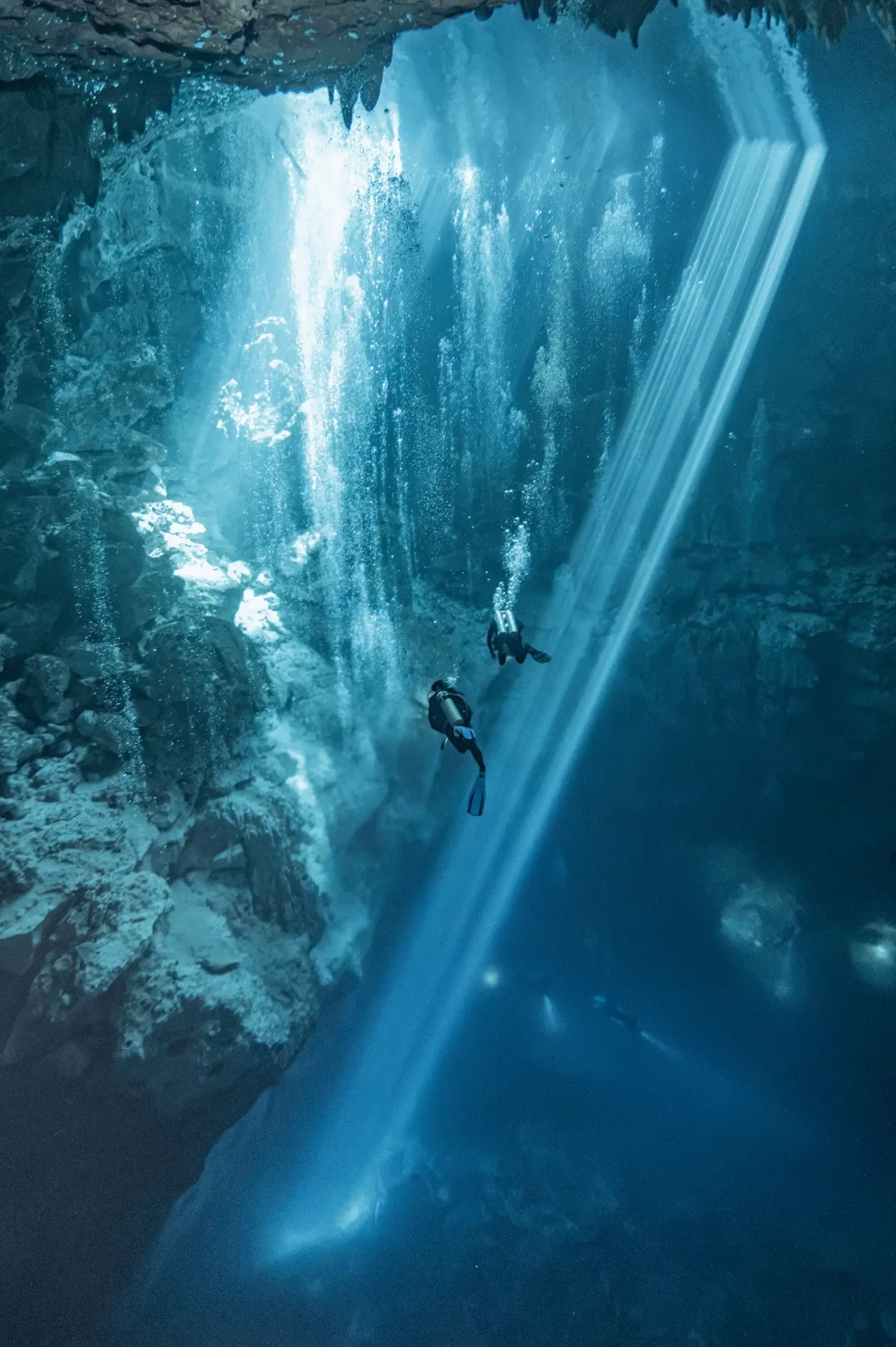Two scuba divers crossing a bright shaft of light inside a blue cenote cavern