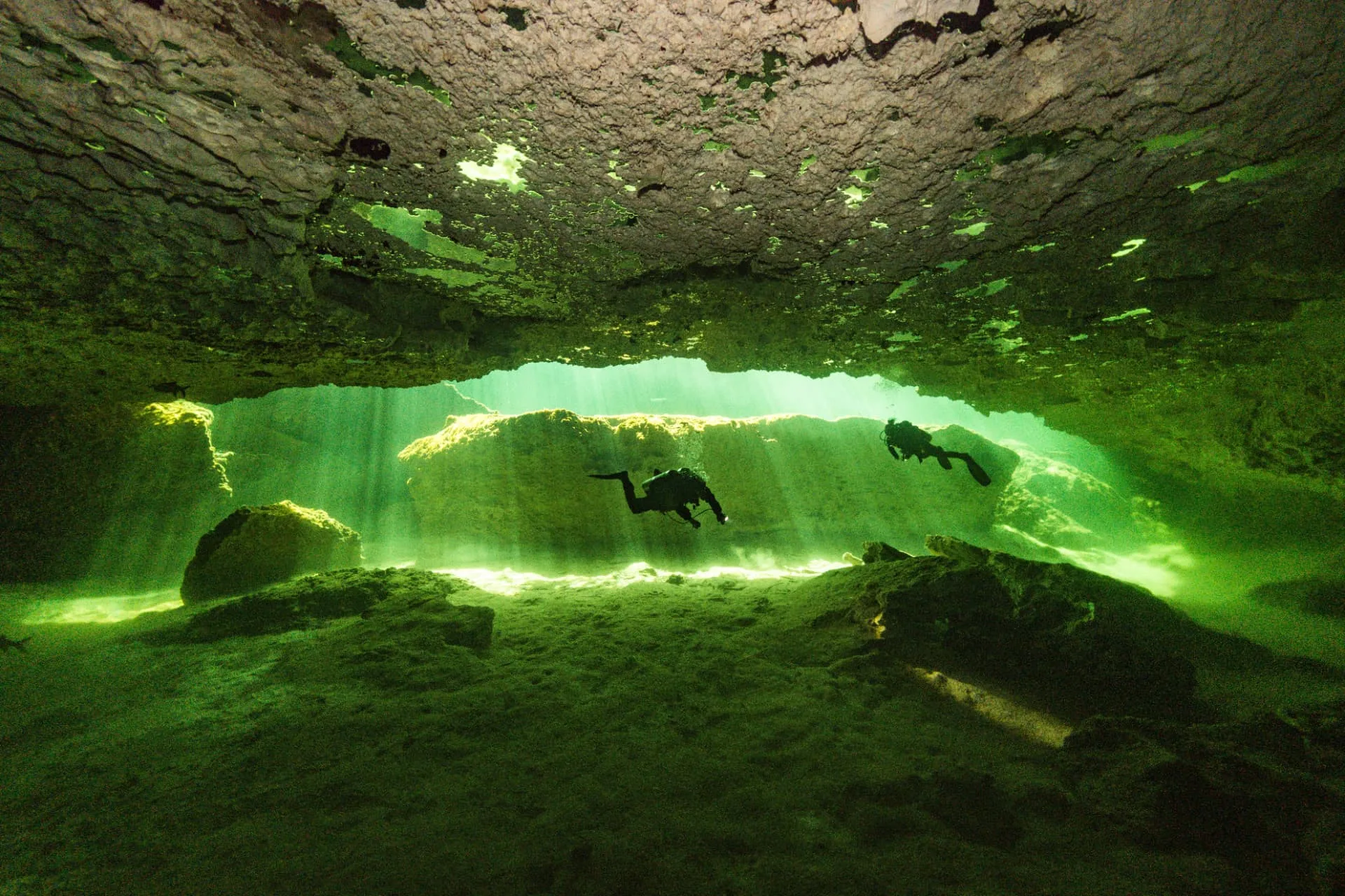Divers swimming together below a rock overhang in calm, clear water