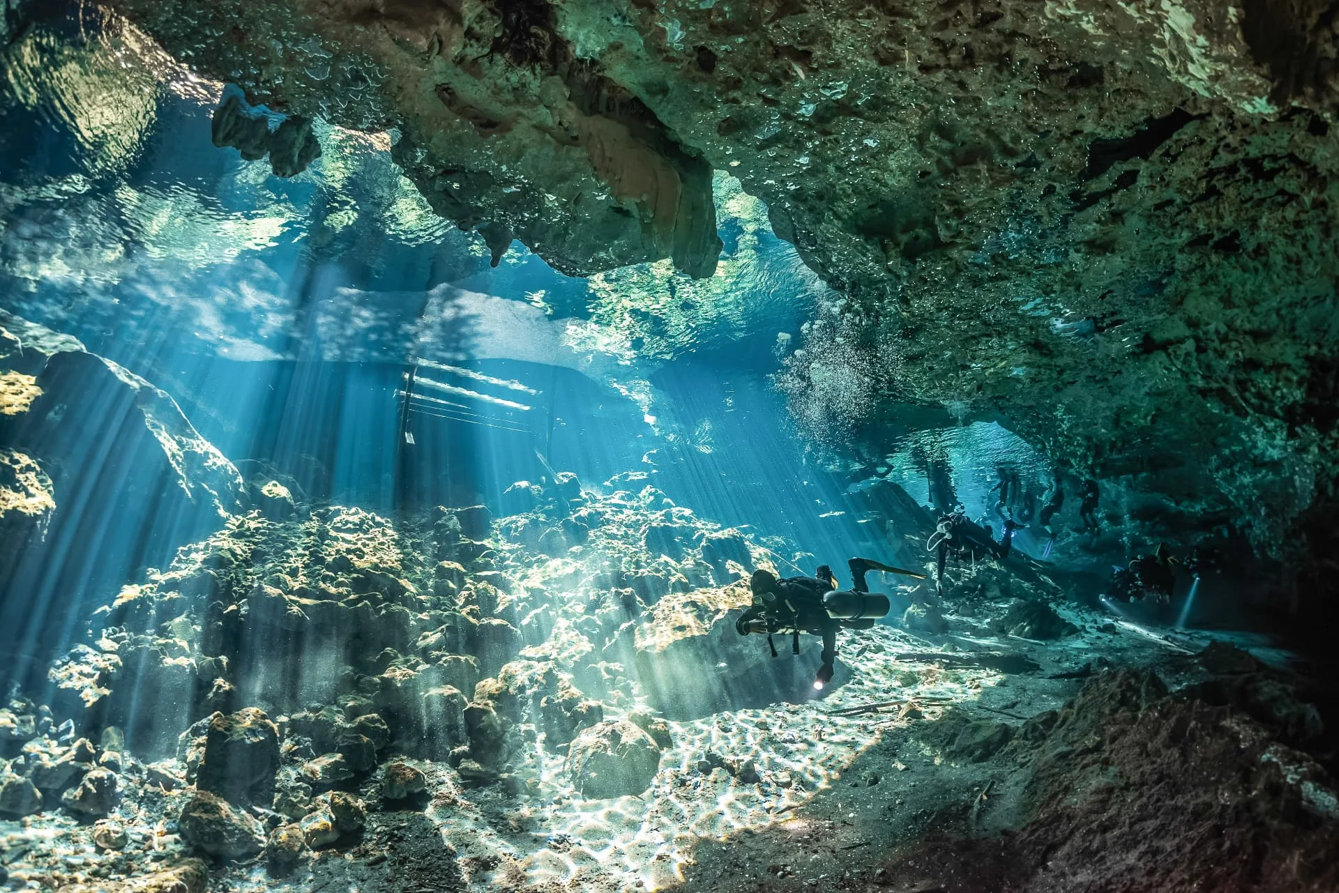 Divers crossing a rocky cavern through strong sunbeams at Nohoch Nah Chich