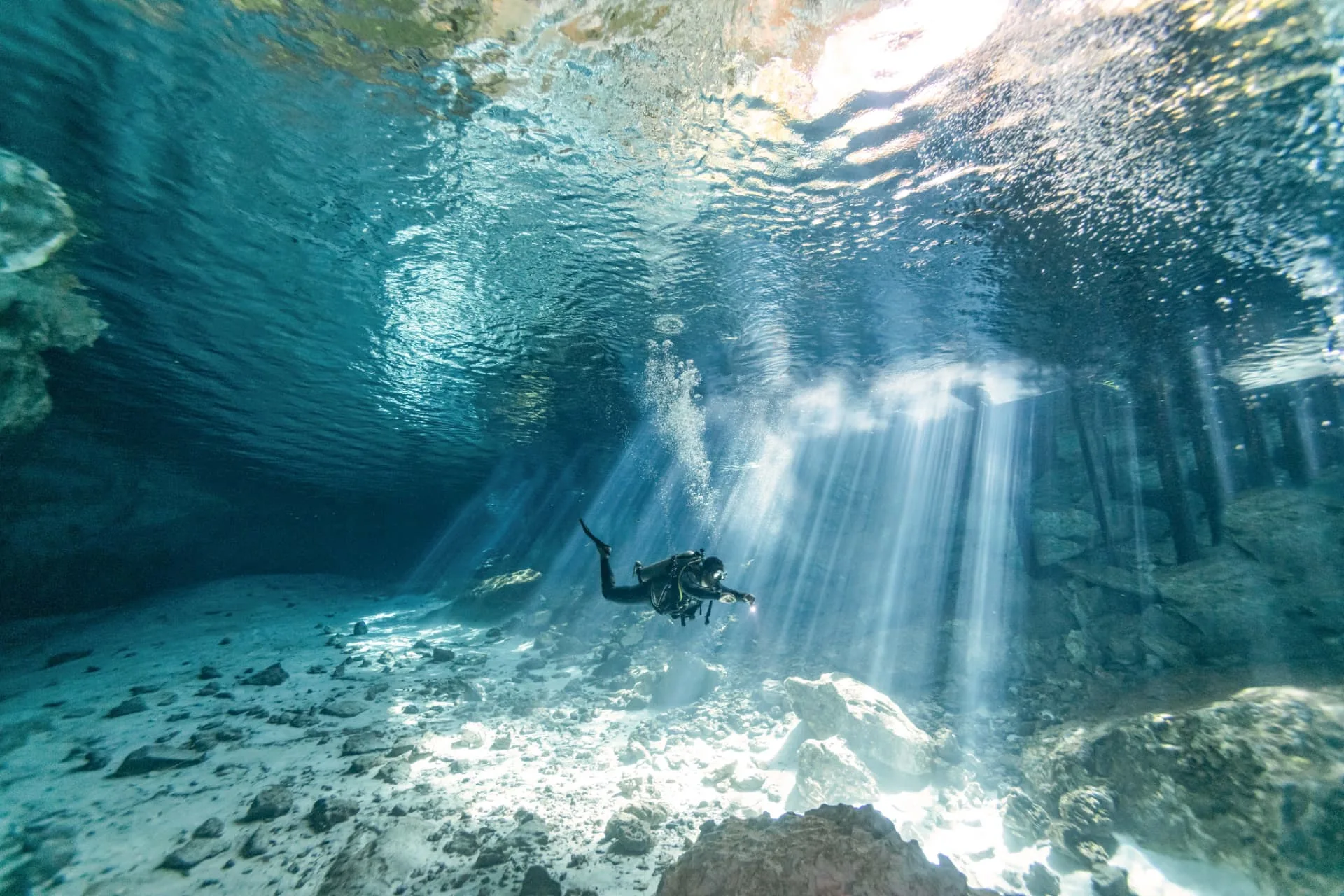 Diver crossing bright sunbeams above the pale rocky floor during a Dos Ojos cavern dive