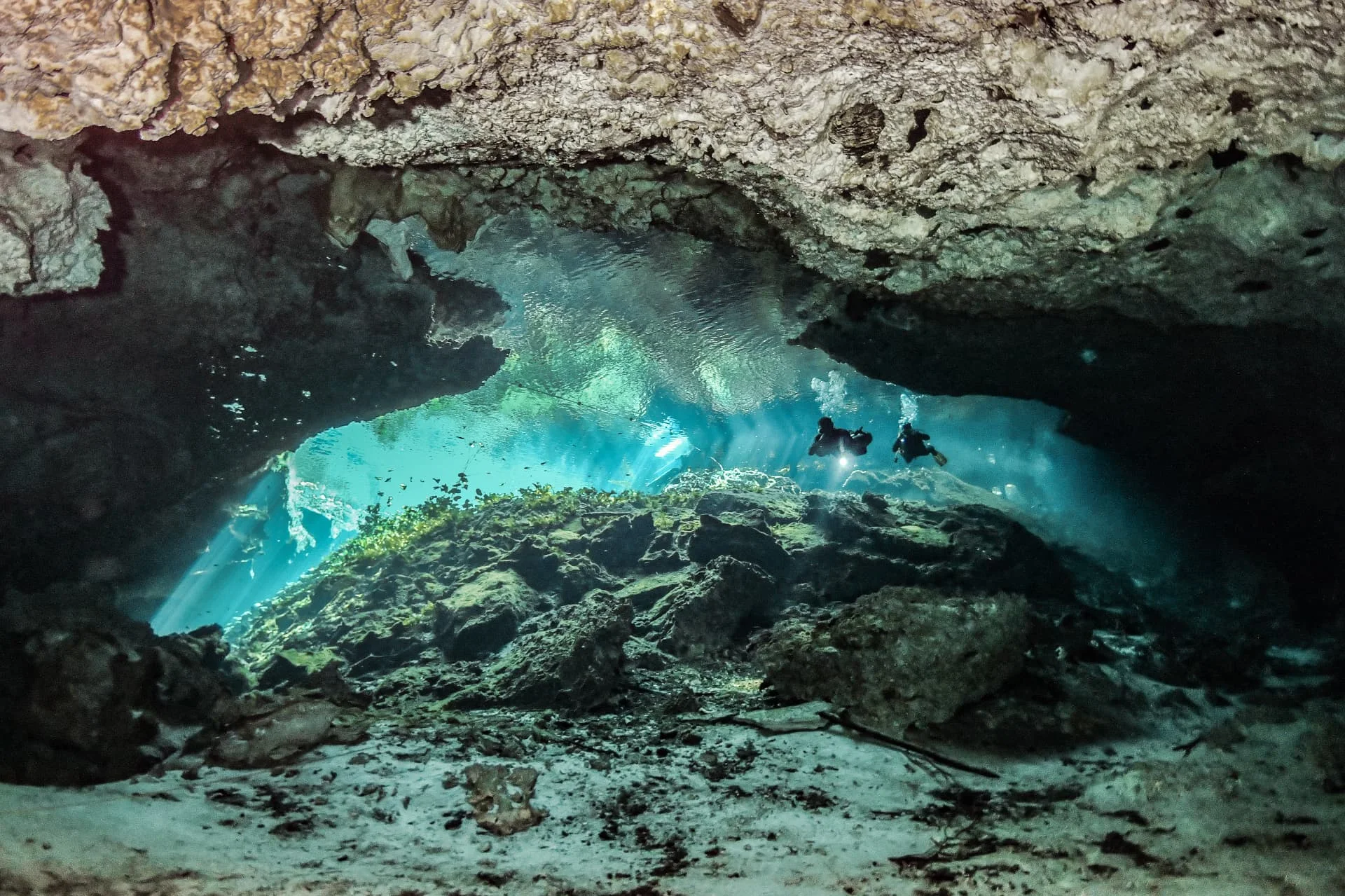 Divers silhouetted below a low rock ceiling near the bright opening at Nicte-Ha